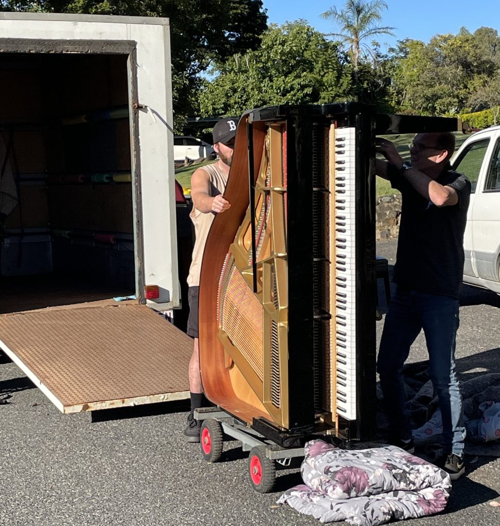 Two men wheel out a black grand piano on a trolley on its side