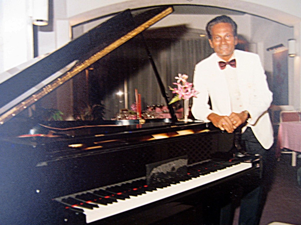 Peter Pragas Malaysian pianist composer standing at a grand piano wearing white tuxedo